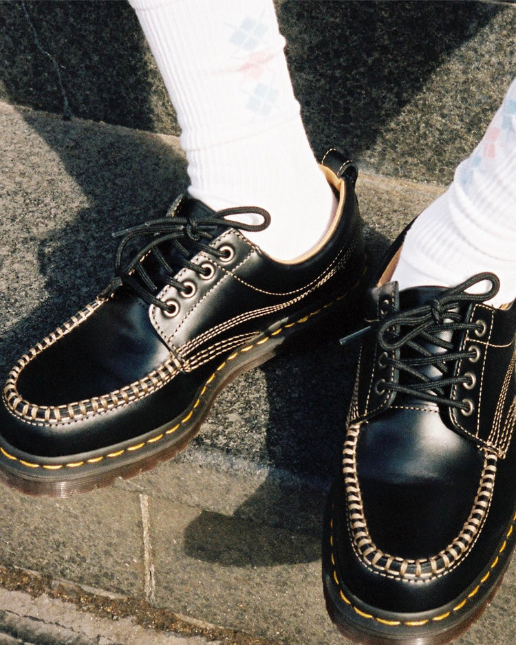 A grainy shot of the Dr. Martens Moc Toe Lowell shoe in black paired with high white socks